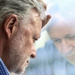 A man looks contemplatively at his reflection in a window