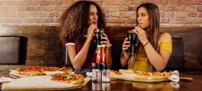 Two women drinking sugar-filled cola in front of unhealthy food