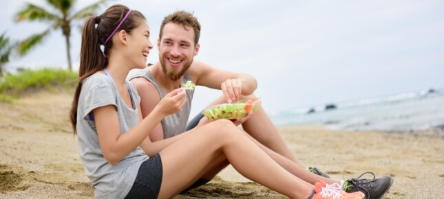 A woman and man eating salad on the beach.