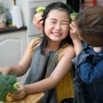 Asian mother holding broccoli against little girl's face.