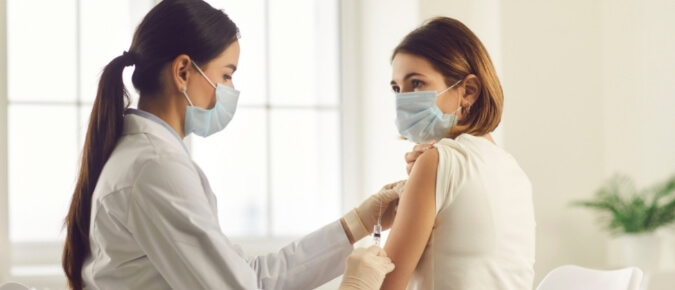 Masked doctor giving vaccination to young woman wearing mask.