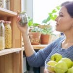 Woman standing in front of her pantry looking at a jar and holding apples.