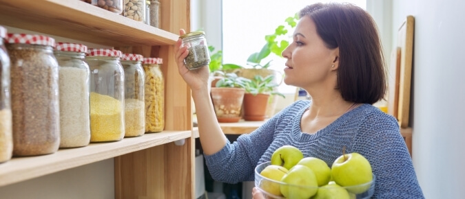 Woman standing in front of her pantry looking at a jar and holding apples.
