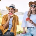 Two women wearing hats and sunglasses eating watermelon outdoors.