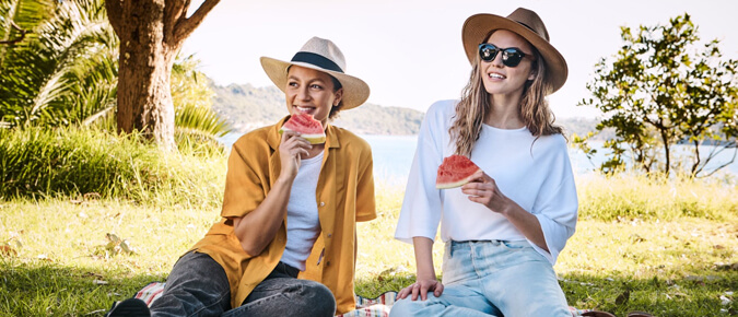 Two women wearing hats and sunglasses eating watermelon outdoors. 