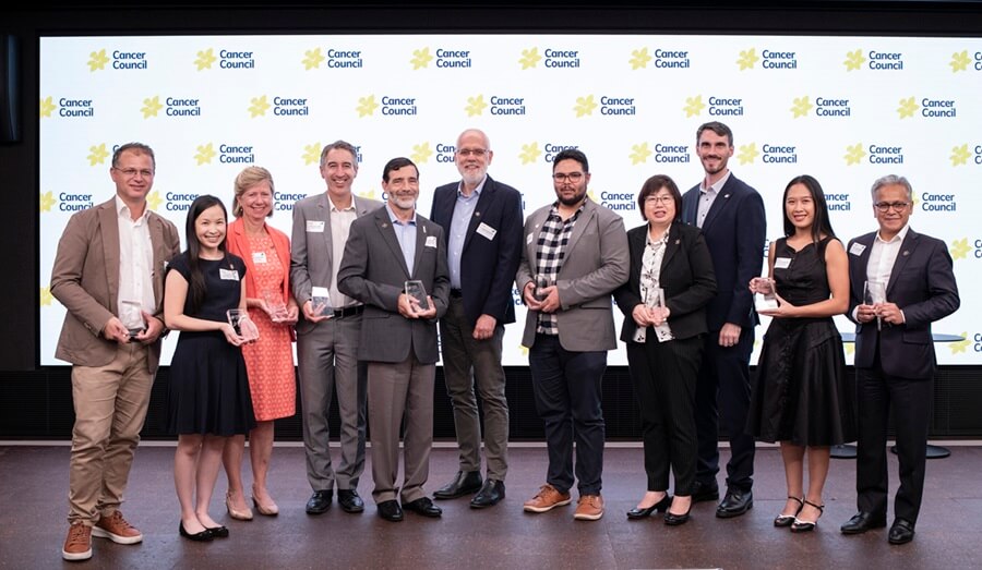 The recipients of the 2025 Cancer Council NSW research awards stand holding their awards in front of a Cancer Council media backdrop.