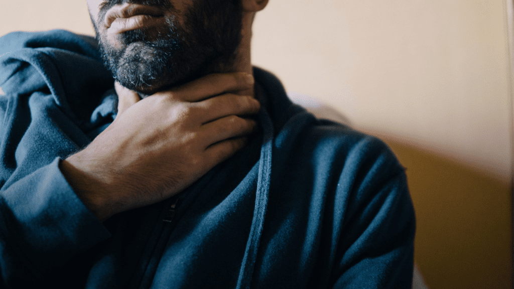 A photo of a man in a hoodie holding his throat, showing discomfort or pain, which can be a symptom of head and neck cancer.