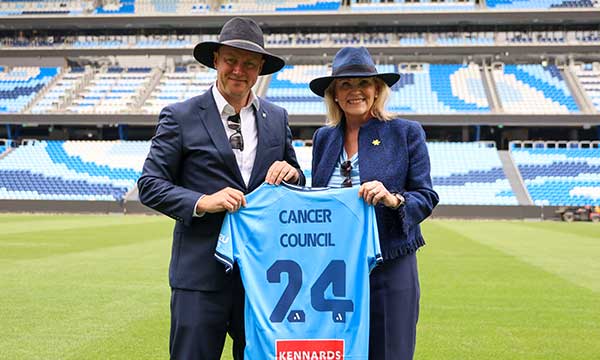 two people hold up a Sydney FC shirt with Cancer Council printed on the back